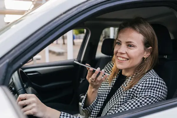 Bilde tatt gjennom åpent vindu på sjåførsiden av en bil, kvinne sitter bak rattet og snakker i en telefon som er på høyttaler, har på seg blazer i grå ruter.