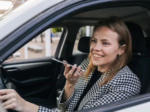 Bilde tatt gjennom åpent vindu på sjåførsiden av en bil, kvinne sitter bak rattet og snakker i en telefon som er på høyttaler, har på seg blazer i grå ruter.
