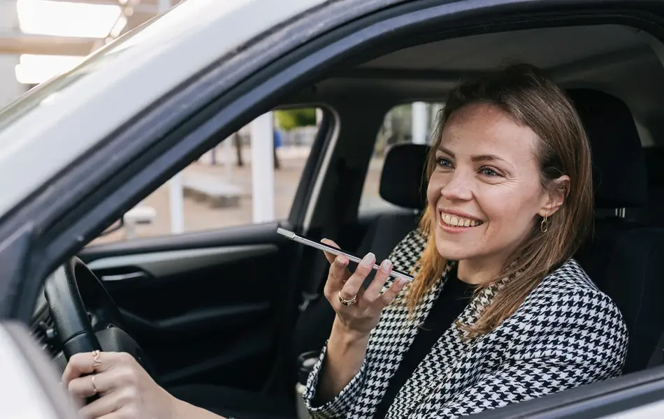 Bilde tatt gjennom åpent vindu på sjåførsiden av en bil, kvinne sitter bak rattet og snakker i en telefon som er på høyttaler, har på seg blazer i grå ruter.