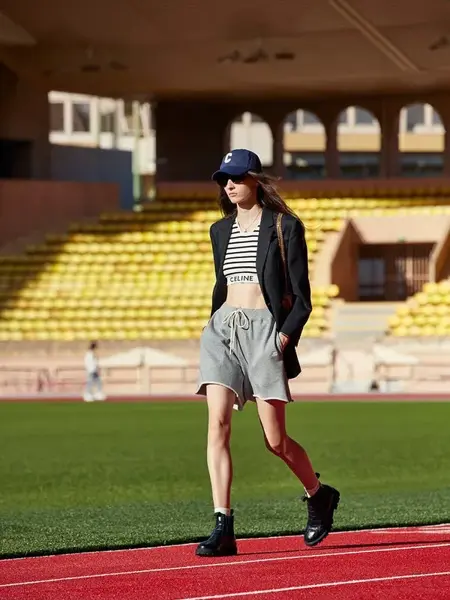 Dame som går ute på en idrettsbane med grå shorts, sort dressjakke, stripete kort magetopp og blå caps