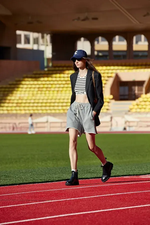 Dame som går ute på en idrettsbane med grå shorts, sort dressjakke, stripete kort magetopp og blå caps