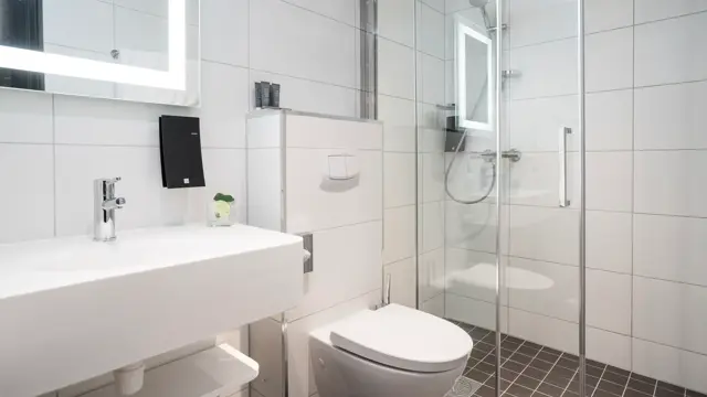 Baderom med god plass, hvitt kakel på vegger og mørkebrunt på gulv. Vegghent toalett, vask og speil med led lys rundt, i superior room på Thon Hotel Polar i Tromsø. 