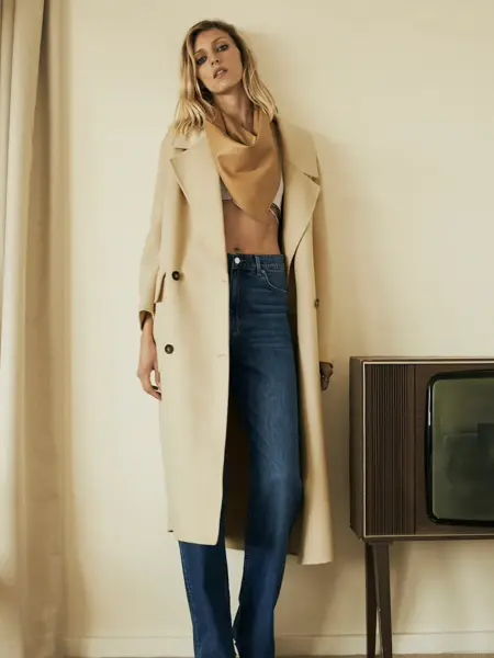 Dame i beige maxikåpe med dongeribukser og skjerf