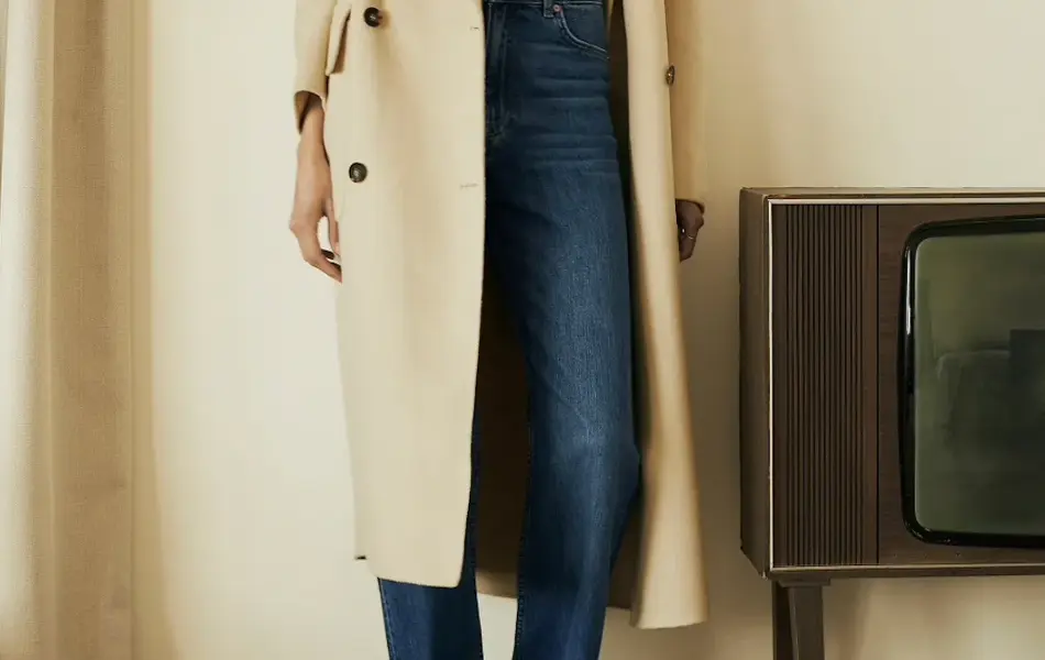 Dame i beige maxikåpe med dongeribukser og skjerf