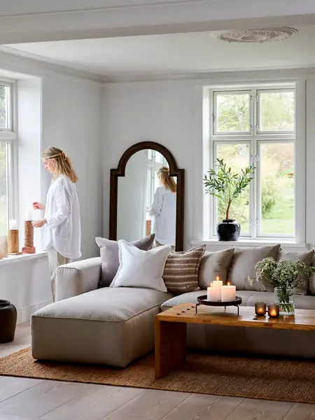 Hvitmalt stue med sofa med puter, sofabord med tente lys, dame står i vinduet og ser ut.