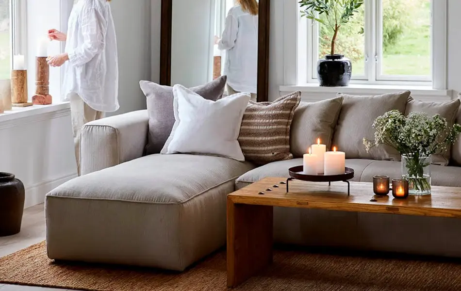 Hvitmalt stue med sofa med puter, sofabord med tente lys, dame står i vinduet og ser ut.