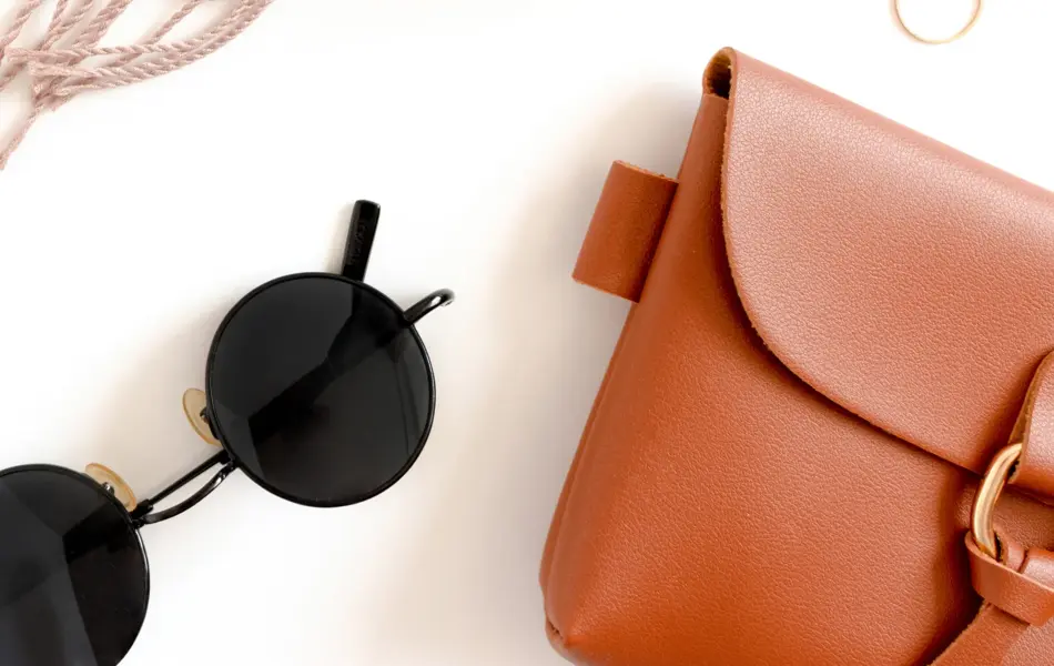 Tilbehør dandert utover en hvit flate. Rosa skjerf, rosa øyenskygge, gullringer, parfymeflaske uten etikett, svarte runde solbriller og brun skinnveske med detaljer i gull. 