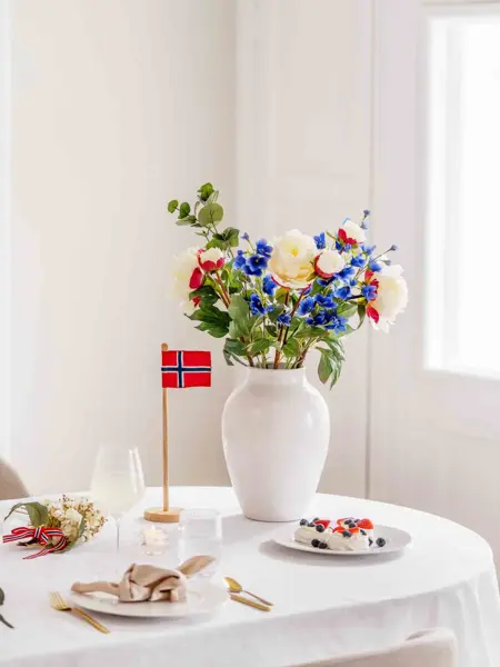 Bord dekket med hvit duk, hvite tallerkner, beige linservietter, vinglass, hvit vase med fargerik bukett, bordflagg.