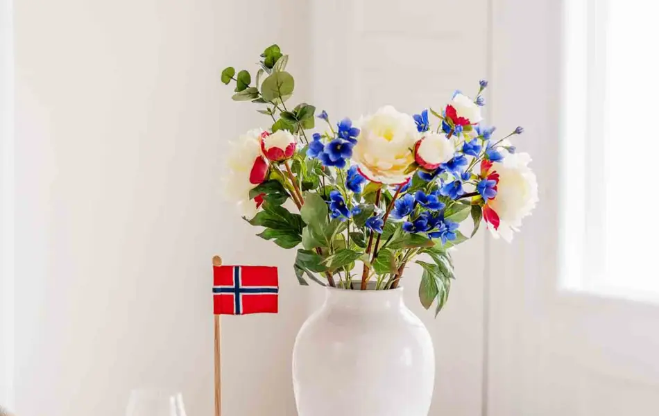 Bord dekket med hvit duk, hvite tallerkner, beige linservietter, vinglass, hvit vase med fargerik bukett, bordflagg.