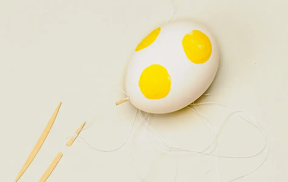 Et hvitt egg med store gule prikker malt på, og tannpirkere og hvit sytråd