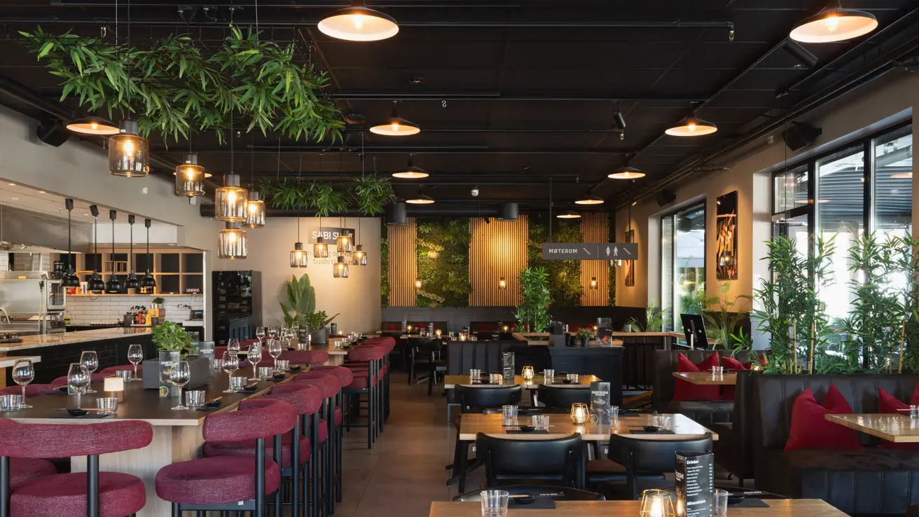 Oversiktsbilde av restauranten med flere bord, bar, grønne planter og hyggelig belysning