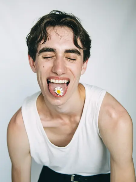 Mann i hvit singlet som geiper til kamera. Han har øynene lukket, og på tunga ligger en hvit blomst.