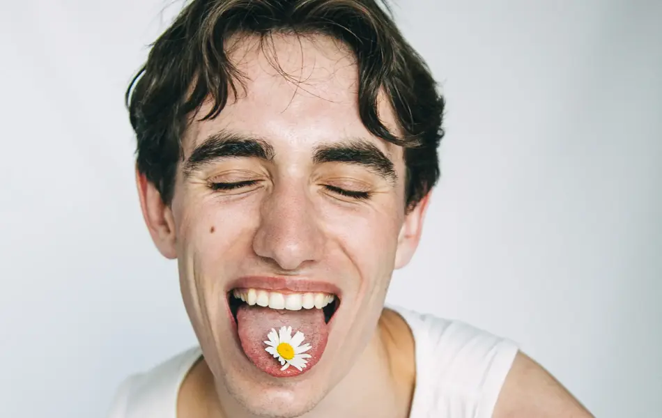 Mann i hvit singlet som geiper til kamera. Han har øynene lukket, og på tunga ligger en hvit blomst.