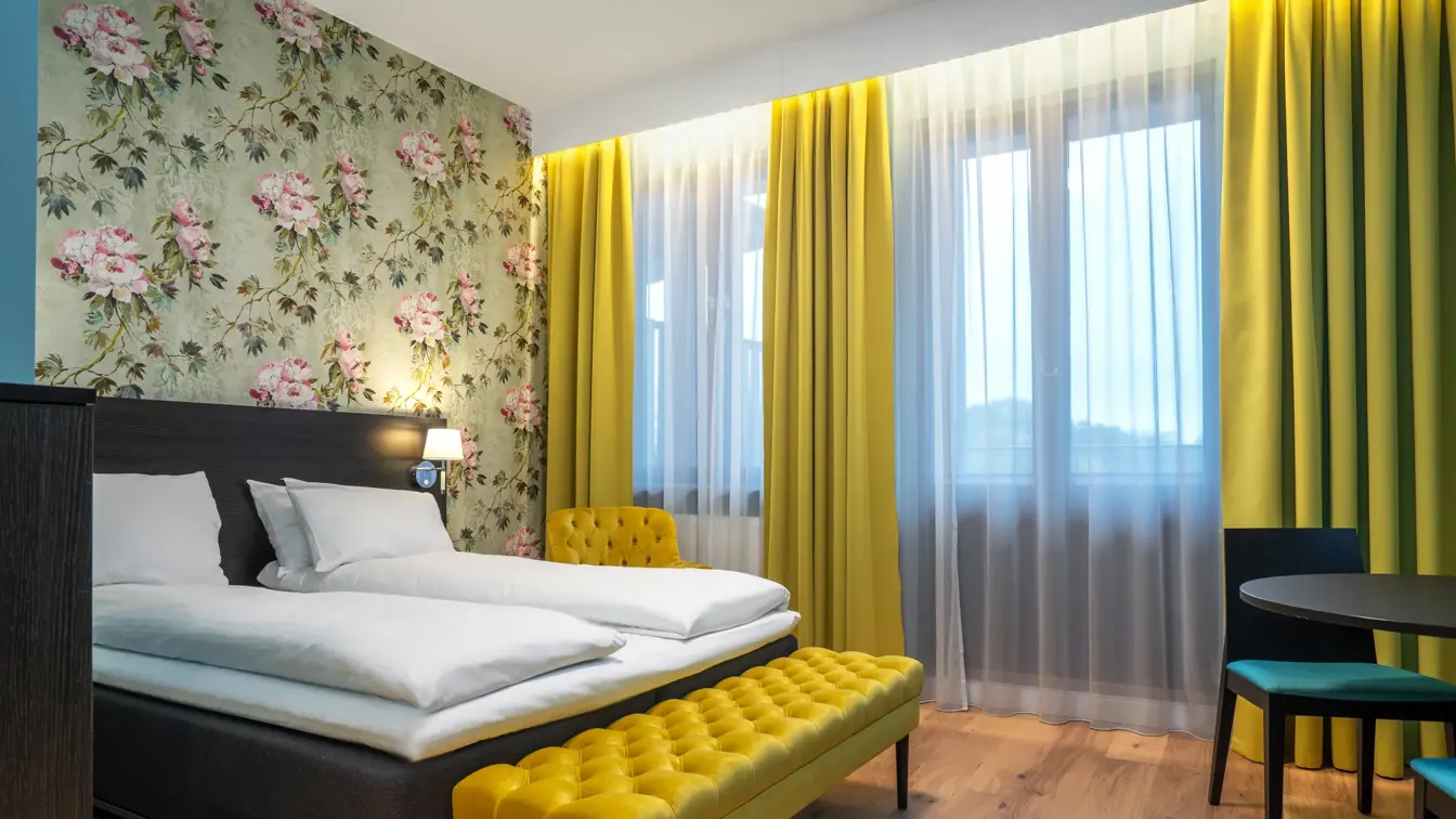 Blomstrete tapetvegg, gule gardiner, gul sofa, gul stol, parkettgulv, dobbeltseng, blå vegger. 