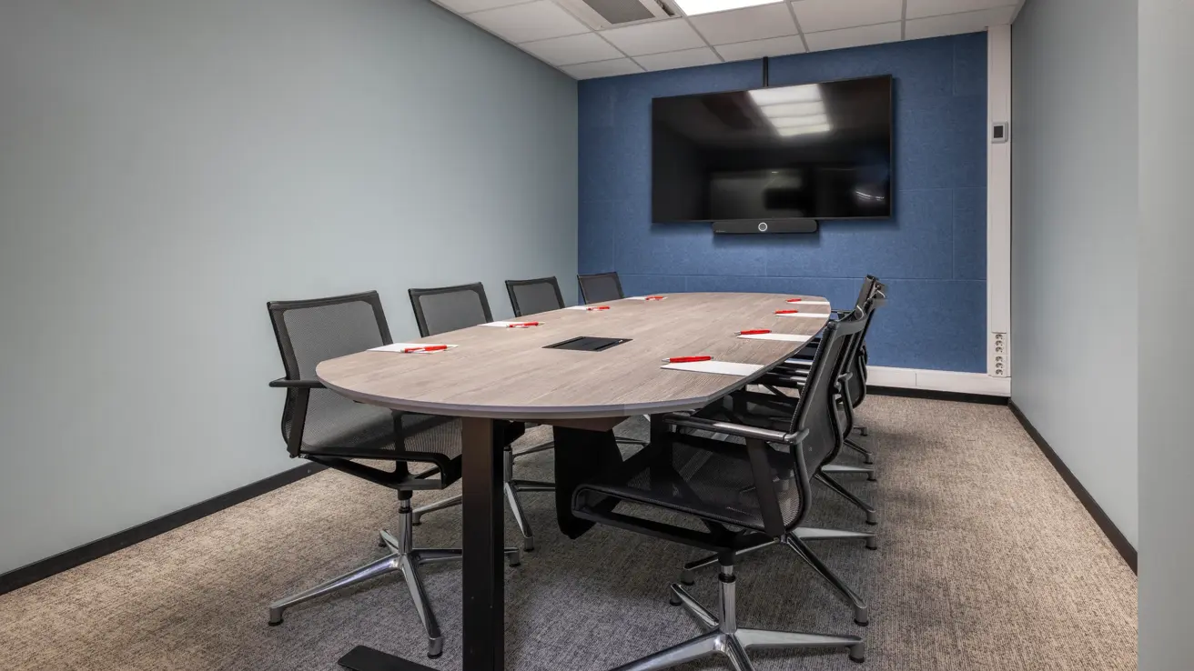 Møterom med styrebords oppsett til 8 personer, blå vegg i enden med sort TV skjerm på veggen.