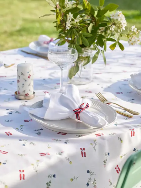 Mønstret duk med hvit tallerken, hvit serviett med flaggbånd rundt, kubbelys og vase med blomster.