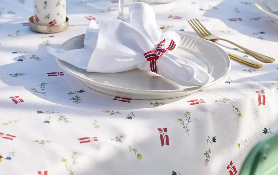 Mønstret duk med hvit tallerken, hvit serviett med flaggbånd rundt, kubbelys og vase med blomster.