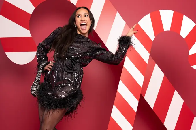 Mona Berntsen i sort festkjole med veske på armen foran en rød vegg med julelpynt.