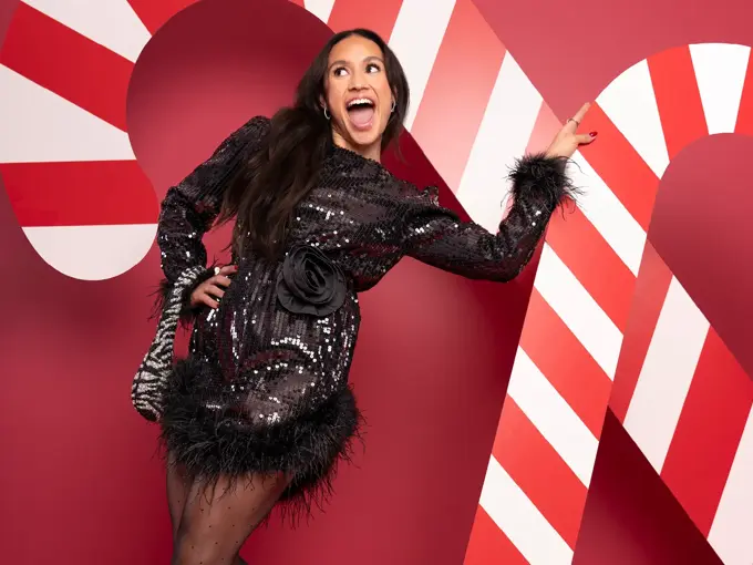 Mona Berntsen i sort festkjole med veske på armen foran en rød vegg med julelpynt.