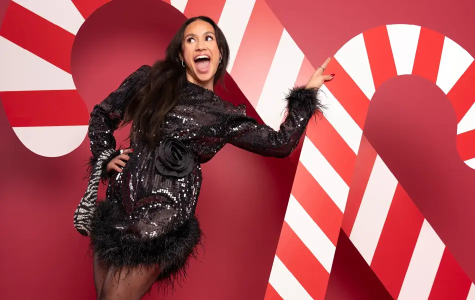 Mona Berntsen i sort festkjole med veske på armen foran en rød vegg med julelpynt.