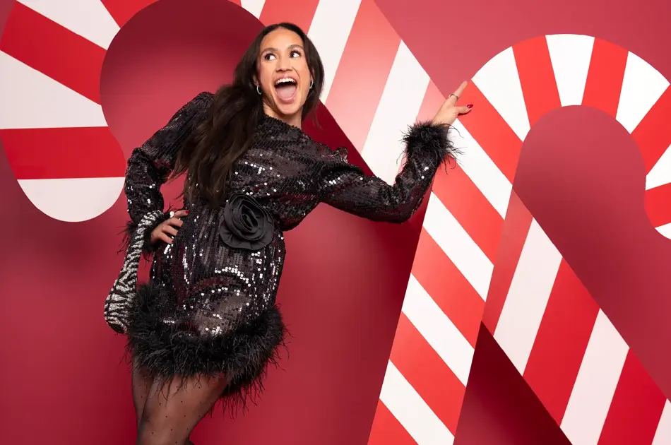 Mona Berntsen i sort festkjole med veske på armen foran en rød vegg med julelpynt.