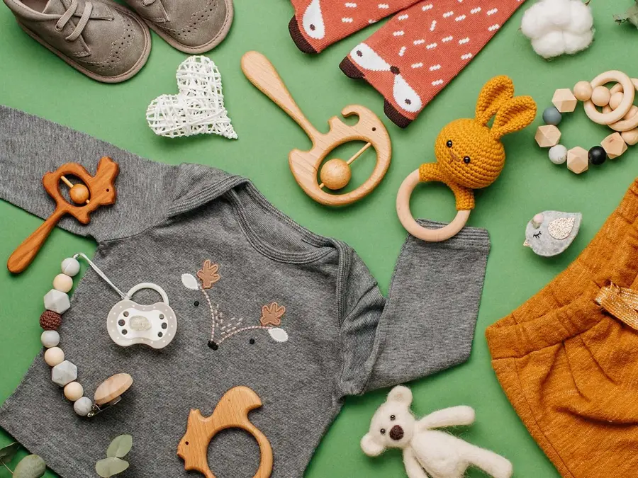 Illustrasjonsfoto til barnebutikk: Barneklær i gult, grått og blått, hvite joggesko, stripete lue, drikkeflaske og bamse på hvit bakgrunn.