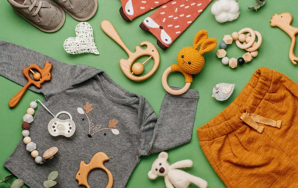 Illustrasjonsfoto til barnebutikk: Barneklær i gult, grått og blått, hvite joggesko, stripete lue, drikkeflaske og bamse på hvit bakgrunn.