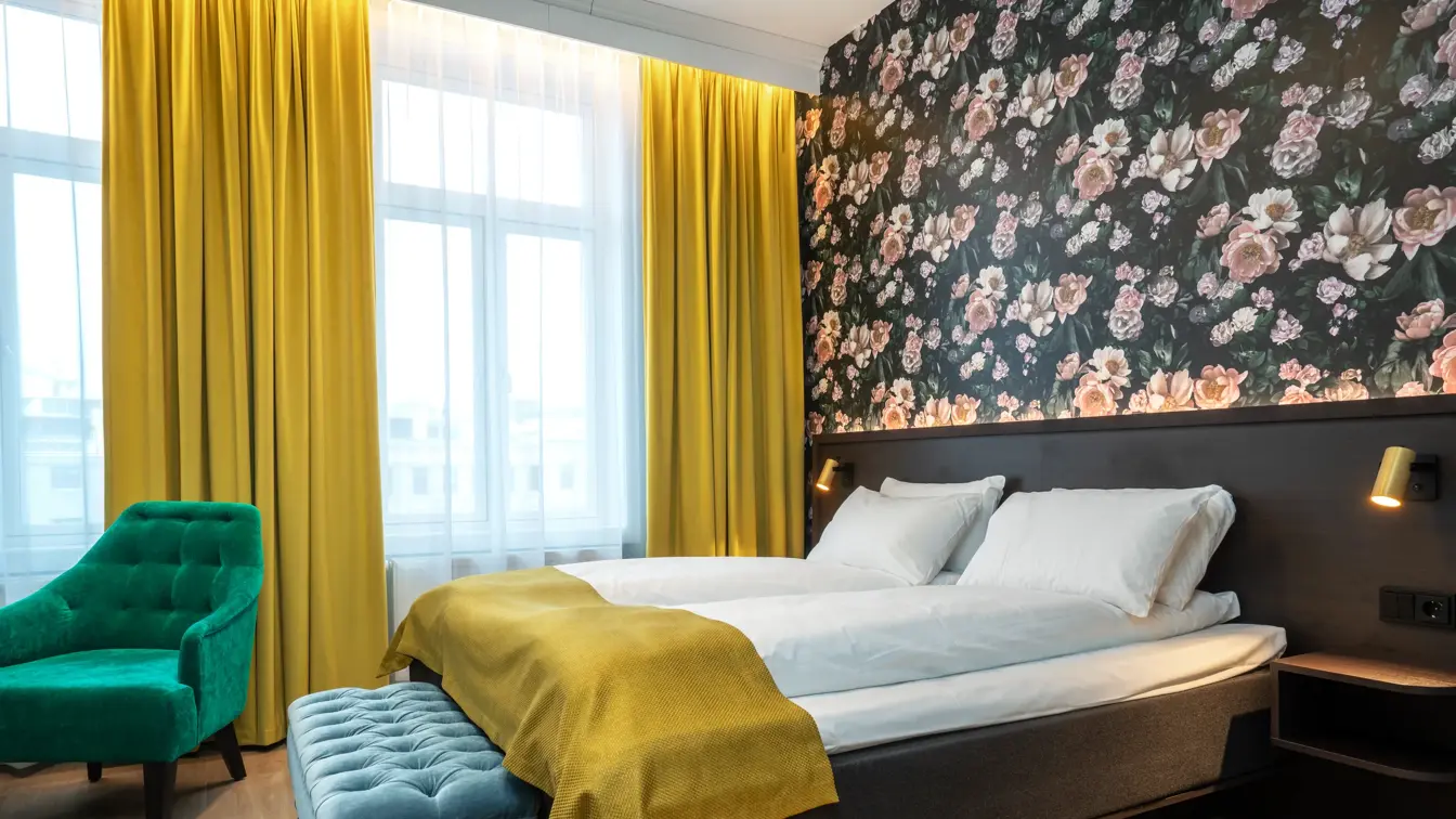Mørk tapet med store lyserosa roser. Store vinduer med gule gardiner fra tak til gulv. Dobbeltseng med gult sengeteppe. Grønn lenestol. I superior rom på Thon Hotel Høyers i Skien.