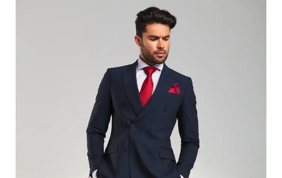 arrogant elegant man in double breasted suit standing with hands in pockets and looks to side on grey studio background