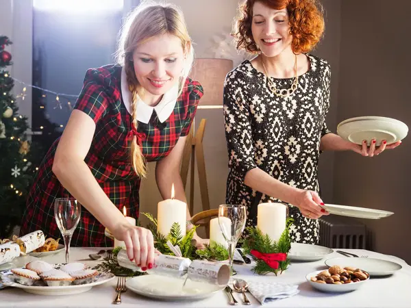 To smilende damer i kjole som dekker på bord med hvite fat, kubbelys og bestikk. Juletre i bakgrunnen.
