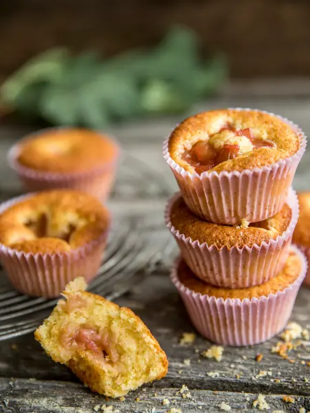 Tre lyse rabarbra muffins stablet oppå hverandre med rosa muffinsform, en halvspist muffins foran, og to i bakgrunnen