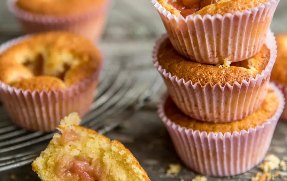 Tre lyse rabarbra muffins stablet oppå hverandre med rosa muffinsform, en halvspist muffins foran, og to i bakgrunnen