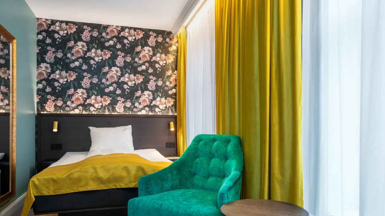 Mørk tapet med store lyserosa roser. Queen size seng meg mørk sengegavl og gult sengeteppe. Vindu med gule gardiner fra tak til gulv. Grønn lenestol og lite mørkt rundt bord. I standard rom queen på Thon Hotel Høyers i Skien.