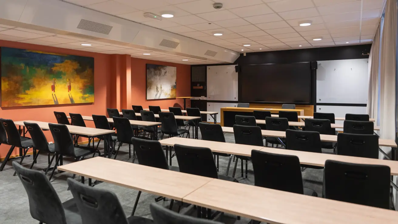 Møterom med klasseroms oppsett, sett bakfra. Svarte stoler, lyse trebord og lys rød farge på veggen.