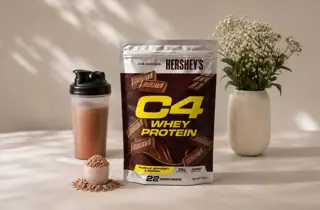 En pose med Whey Protein fra C4 står ved siden av en shaker med drikke i og en vase med blomster.
