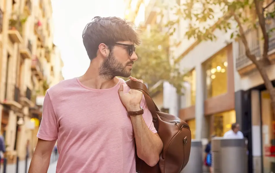 Mann med rosa t-skjorte og brun skinnbag over skulderen poserer på gate utendørs.