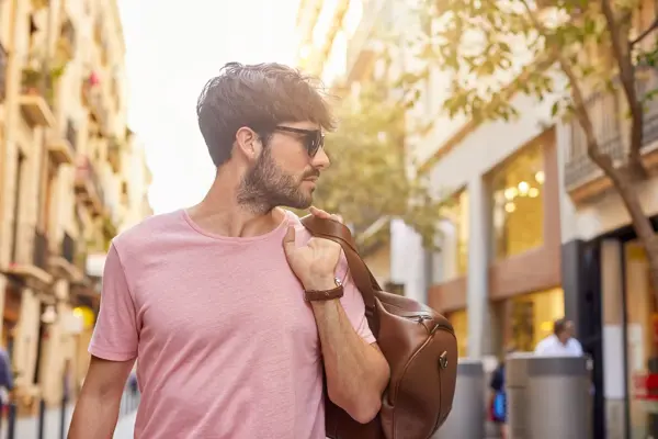 Mann med rosa t-skjorte og brun skinnbag over skulderen poserer på gate utendørs.