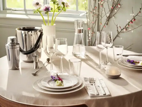 rundt bord med hvit duk, hvitt service, glass og vinkjøler. Vase med blomster.