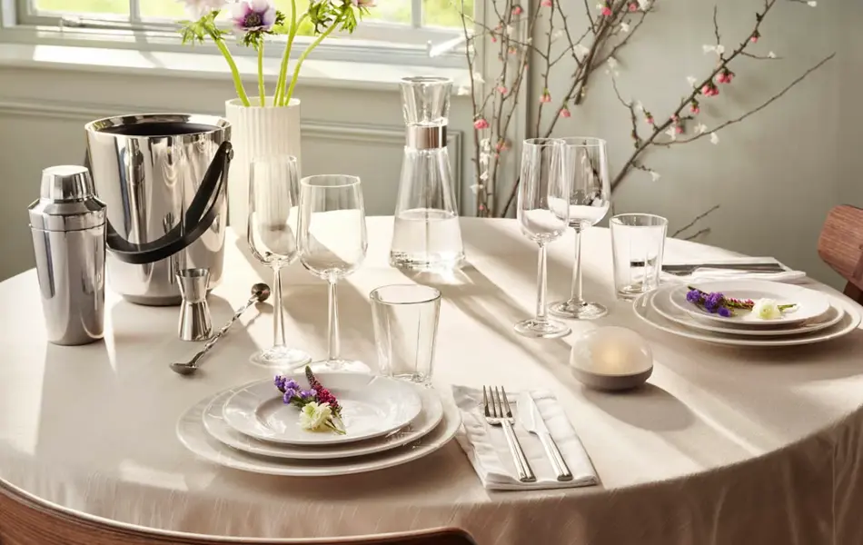 rundt bord med hvit duk, hvitt service, glass og vinkjøler. Vase med blomster.