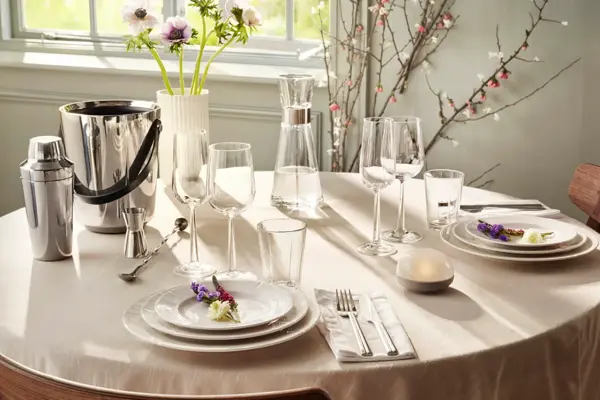 rundt bord med hvit duk, hvitt service, glass og vinkjøler. Vase med blomster.