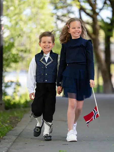 Liten gutt i mørk blå festdrakt holder en jente i hånden med mørk blå kjole, hvite sneakers og holder et flagg.