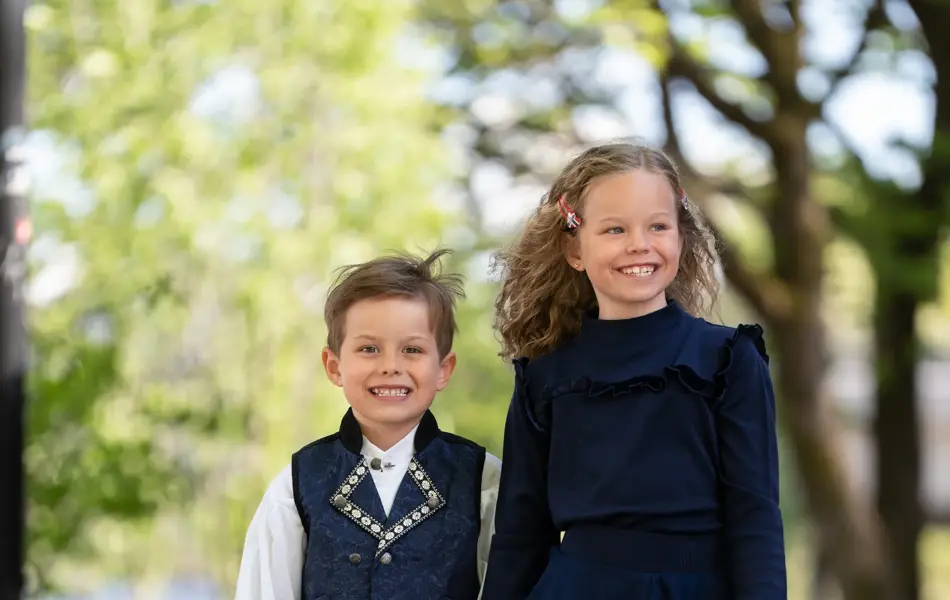 Liten gutt i mørk blå festdrakt holder en jente i hånden med mørk blå kjole, hvite sneakers og holder et flagg.