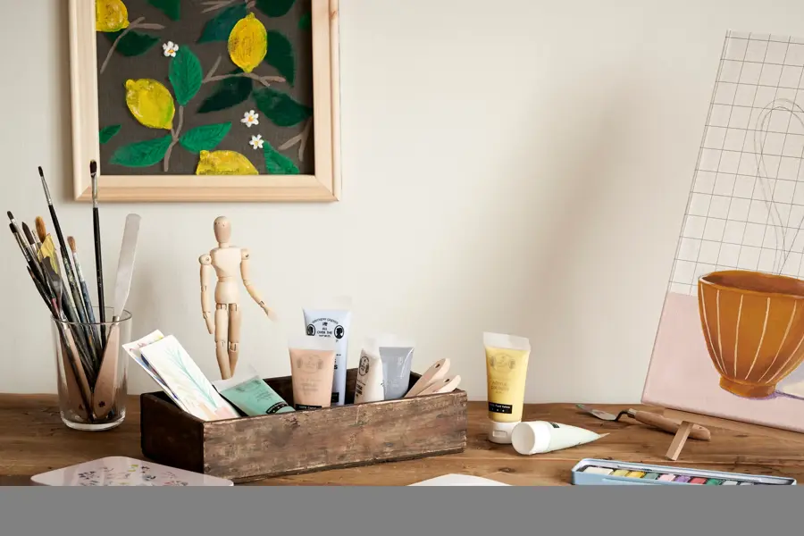 Pult med malepensler og maling, dekormannfigur, malingsbilde på vegg og bordmaleri på lærret og staffeli som alt selges på Søstrene Grene