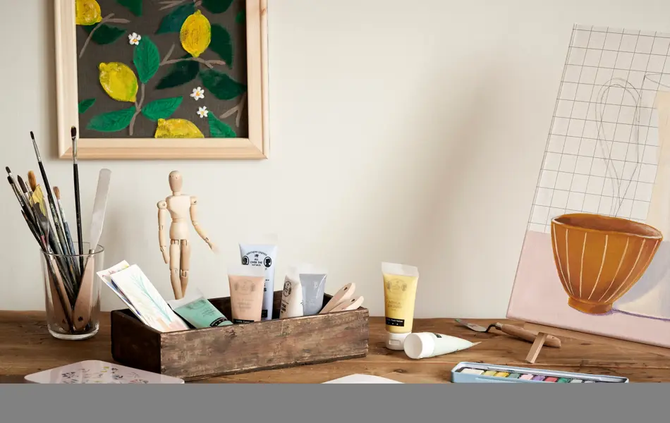 Pult med malepensler og maling, dekormannfigur, malingsbilde på vegg og bordmaleri på lærret og staffeli som alt selges på Søstrene Grene