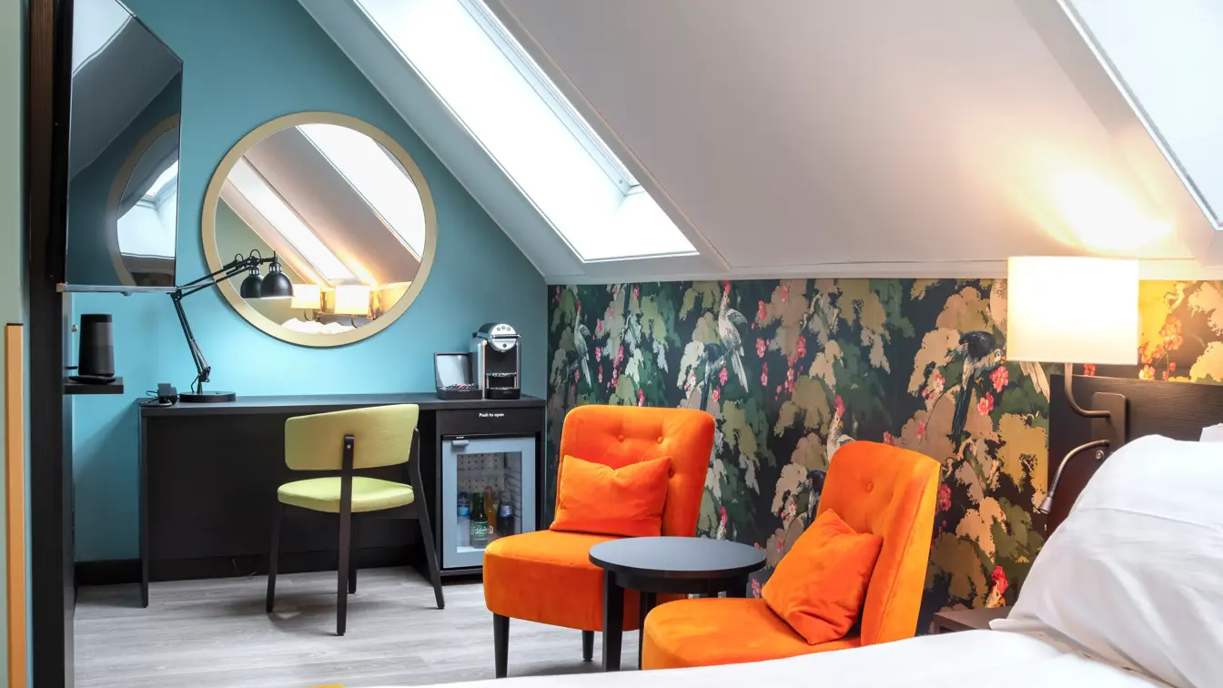 Hvit, turkis og blomstrete tapetvegger, to takvinduer i skråtak, to oransje stoler, avlastningsbord, svart skrivebord med integrert minibar, gul stol, lampe, rundt speil på vegg, dobbeltseng, TV hengende på vegg.