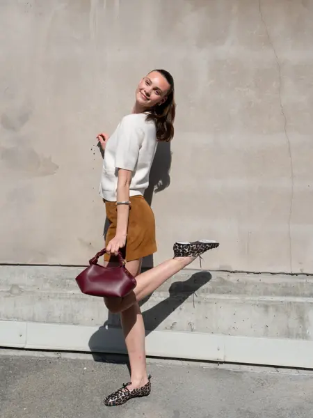 Ung dame i hvit kortermet strikketopp, brun semsket kort skjørt, burgunder veske og flate sko i leopardmønster