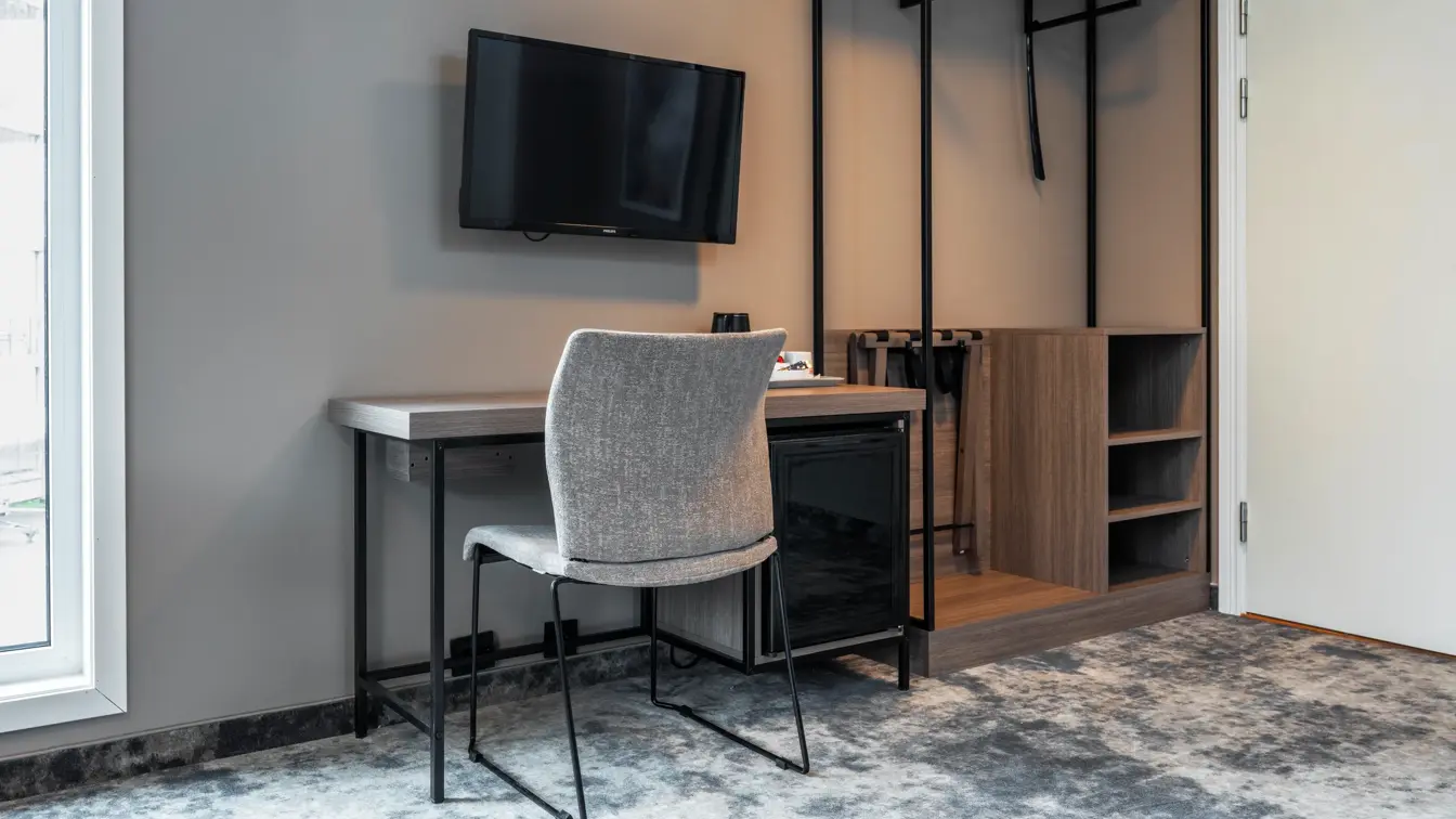 Business rom med skrivebord og tilhørende stol, vegghengt TV, teppegulv og åpen garderobe.