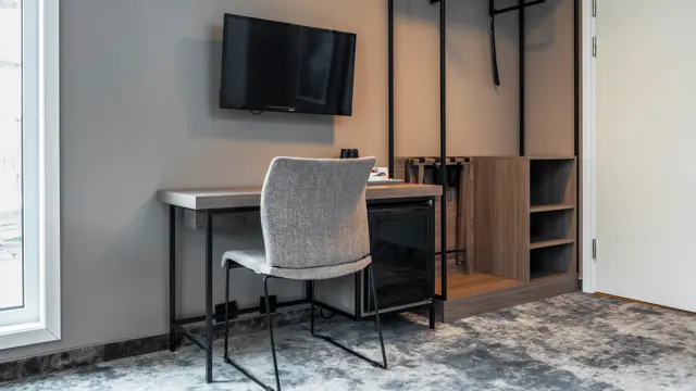 Business rom med skrivebord og tilhørende stol, vegghengt TV, teppegulv og åpen garderobe.