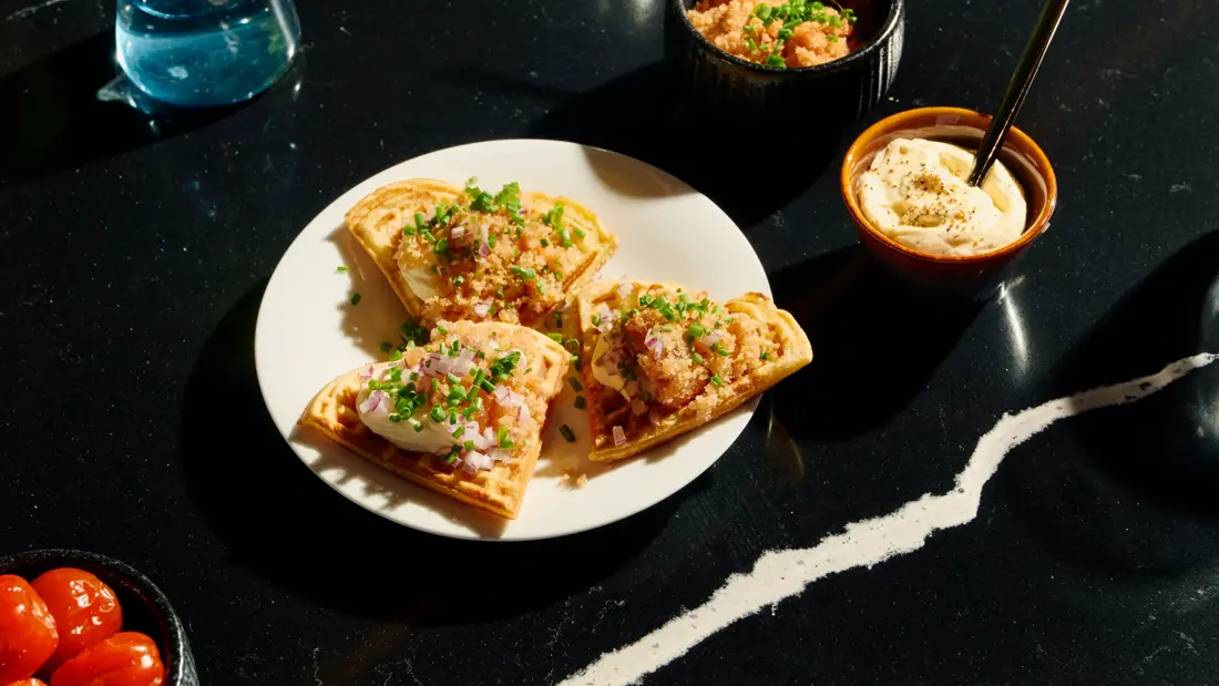 En tallerken med tre hjerteformede vaffelbiter toppet med fiskerogn, rømme, hakket rødløk og gressløk. Ved siden av er to små skåler med ekstra rogn og rømme.