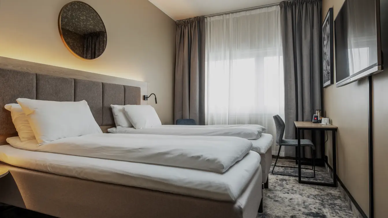 Dobbeltrom med twin senger, hvitt sengetøy, vegghengt TV, gulvteppe, lange grå gardiner og stort vindu i enden av rommet.